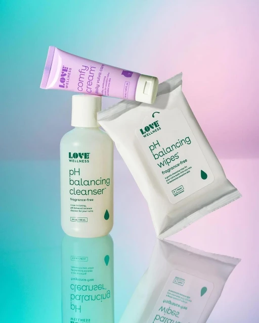 Close-up shot of Love Wellness brand products including pH balancing cleanser, comfy cream, and pH balancing wipes on reflective surface.