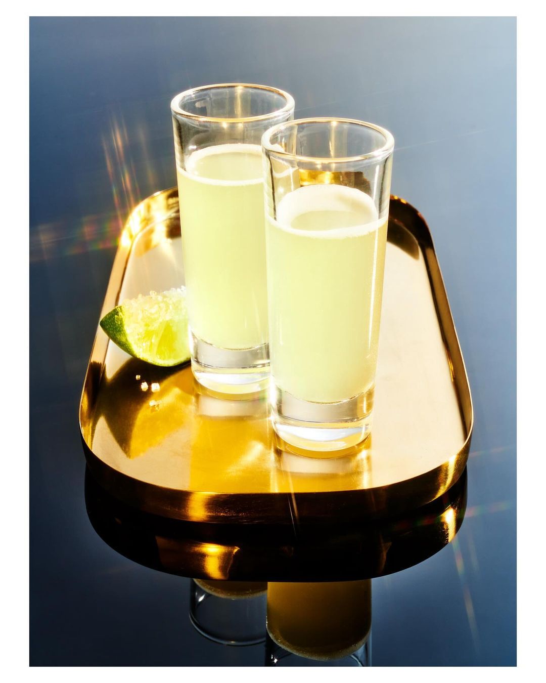 Two shot glasses of lime-colored liquid and a lime wedge sit on a gold serving tray against a gradient dark blue background.
