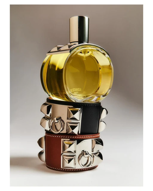 Close-up studio shot of a round Hermès perfume bottle stacked on two studded leather bracelets, product photography, warm lighting.