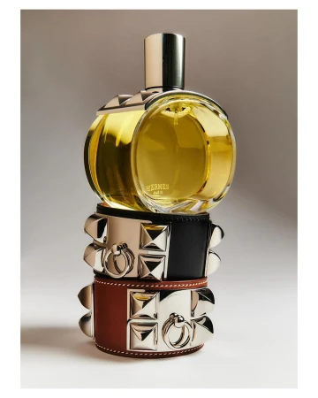 Close-up studio shot of a round Hermès perfume bottle stacked on two studded leather bracelets, product photography, warm lighting.