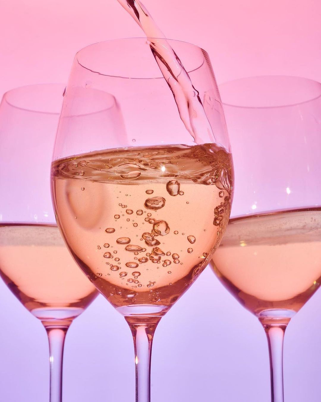 Three wine glasses filled with a white wine, one glass is being poured into, pink and purple color background, close up view.