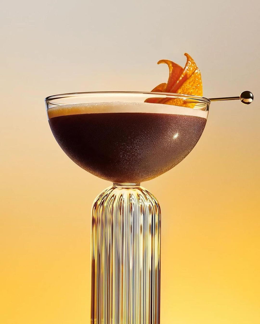 An elegant coupe glass filled with a dark cocktail, garnished with an orange peel twist, set against a gradient yellow background.