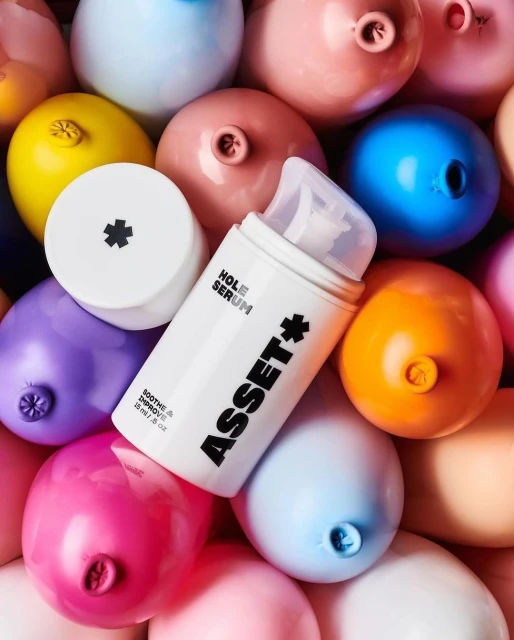 Cosmetic serum bottle, 'Asset + Hole Serum,' surrounded by colorful balloons in a close-up studio product shot.