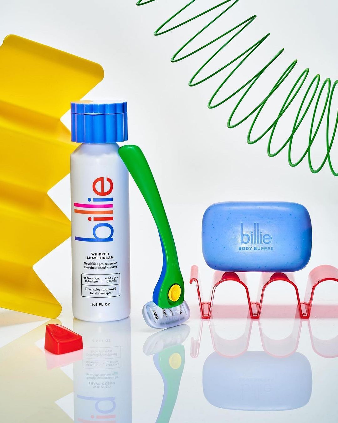 A close-up product shot of Billie brand items including a bottle of shave cream, a razor, and a body buffer against a white background.