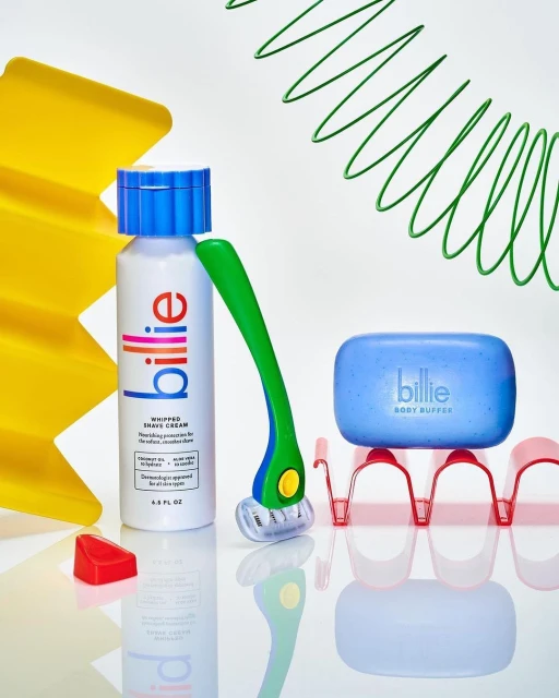 A close-up product shot of Billie brand items including a bottle of shave cream, a razor, and a body buffer against a white background.