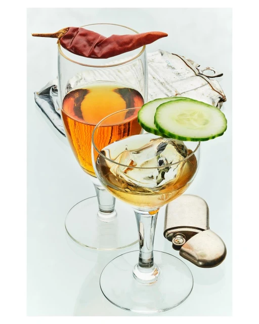 Still life of two stemmed cocktail glasses with liquor, one topped with red chili pepper, cucumber slices and a metallic flask.