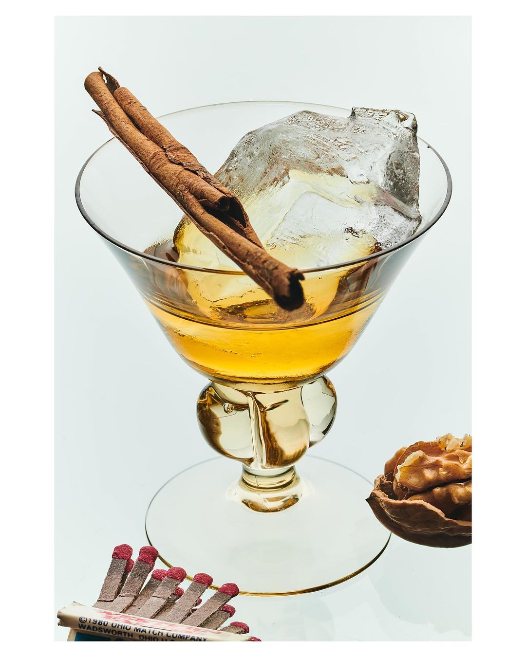 Cocktail in a stemmed glass with ice and cinnamon sticks on a white background, alongside a matchbox and walnut.