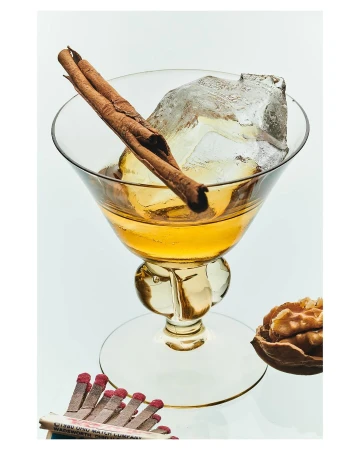 Cocktail in a stemmed glass with ice and cinnamon sticks on a white background, alongside a matchbox and walnut.