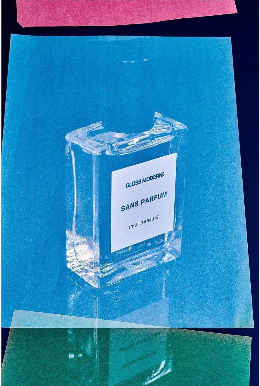 Clear glass bottle of 'Gloss Moderne Sans Parfum L'Huile Beaute' on a blue background with pink and green paper elements.