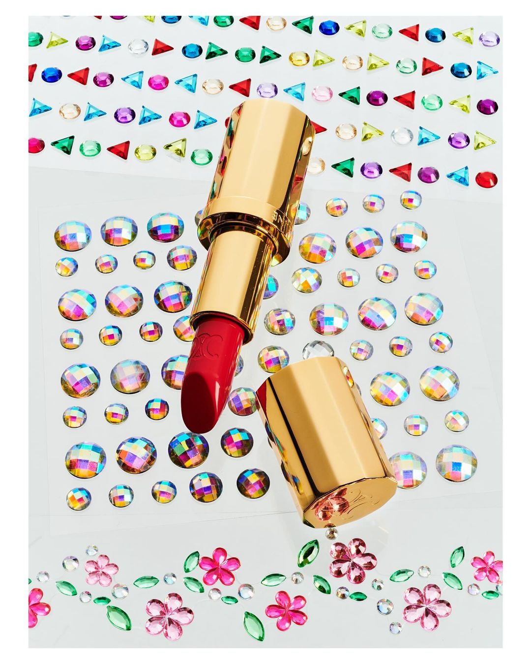 High-angle studio shot of a red lipstick tube with a gold case, surrounded by multicolored rhinestone embellishments on a white surface.