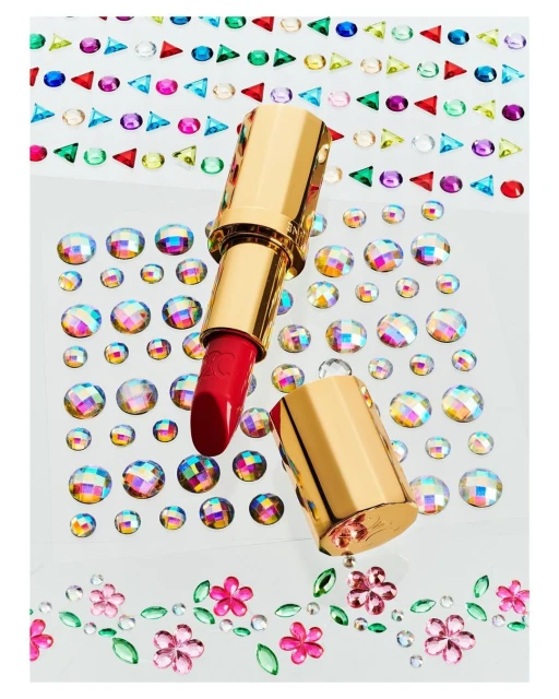 High-angle studio shot of a red lipstick tube with a gold case, surrounded by multicolored rhinestone embellishments on a white surface.