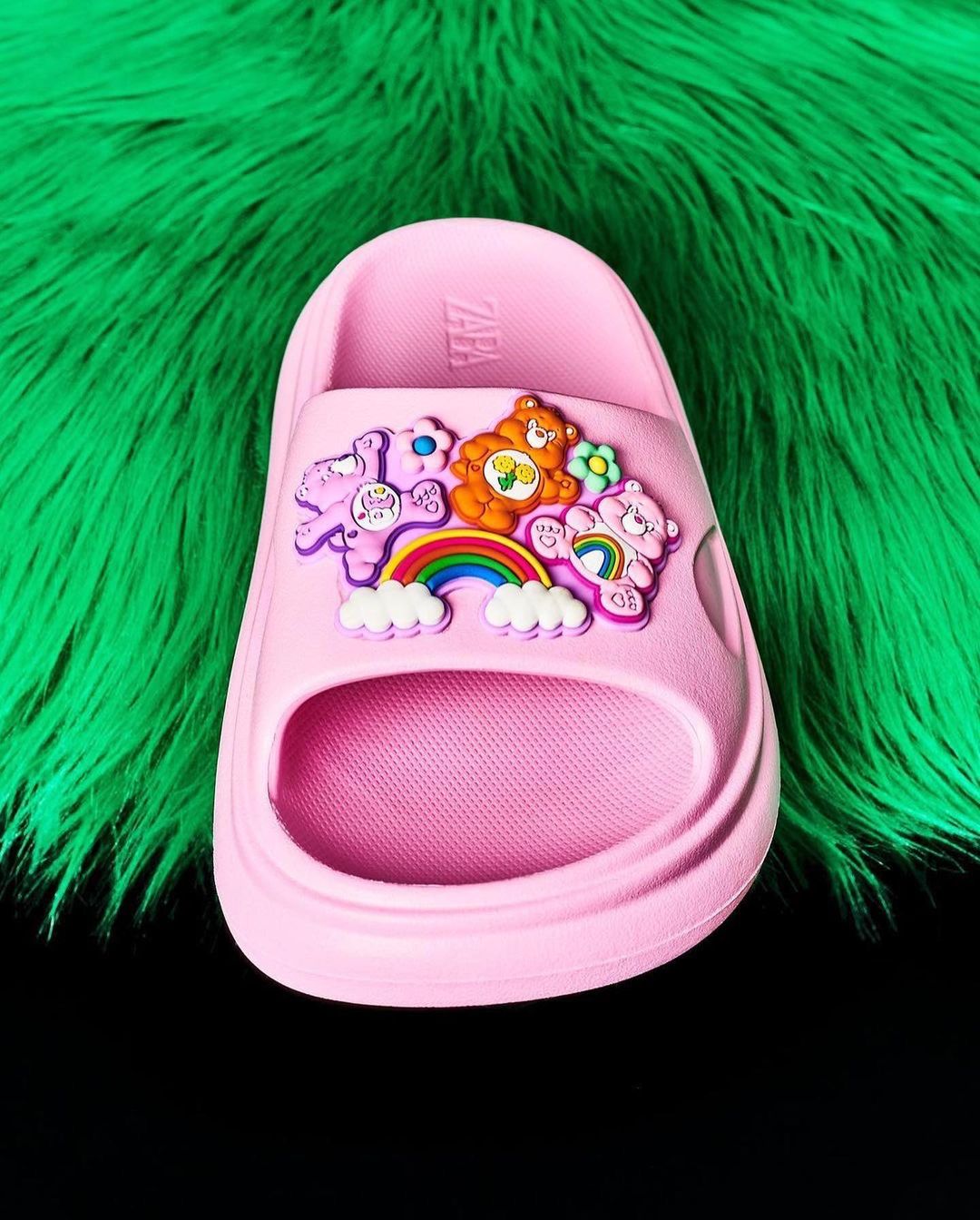 Pink rubber sandal featuring a Care Bears design, set against a vibrant green faux fur and a contrasting black background.
