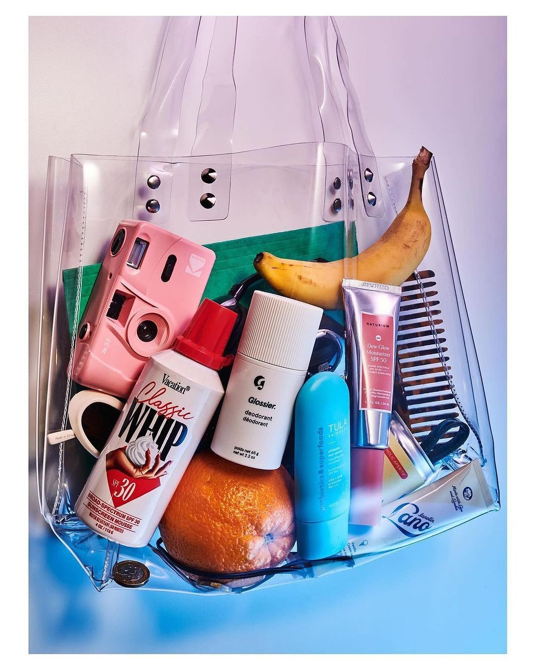 Clear tote bag filled with a camera, sunscreen, deodorant, banana, orange, and comb, against a white background.