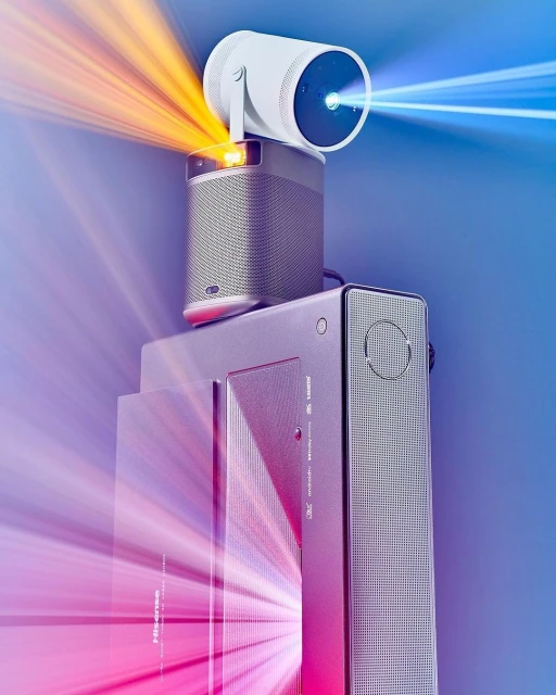 Close-up of a white XGIMI projector and gray soundbar, stacked on top of a Hisense speaker, with colorful light rays in the background.