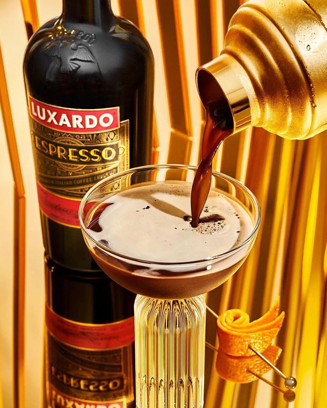 Espresso martini drink being poured into a cocktail glass next to a bottle of Luxardo espresso liqueur with a gold background.