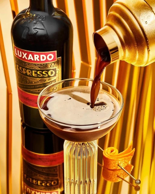 Espresso martini drink being poured into a cocktail glass next to a bottle of Luxardo espresso liqueur with a gold background.