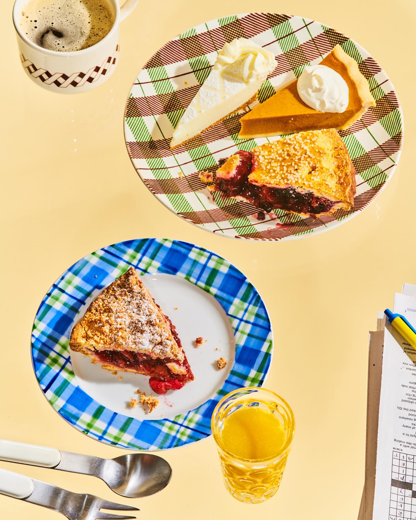 Overhead view of two plates with slices of pie and cake, accompanied by a cup of coffee, a glass of juice, and a crossword puzzle.
