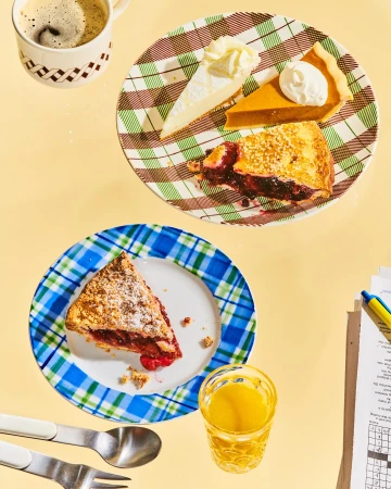 Overhead view of two plates with slices of pie and cake, accompanied by a cup of coffee, a glass of juice, and a crossword puzzle.