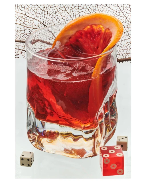 Close-up of a cocktail in a clear glass with a blood orange slice, accompanied by a pair of dice on a reflective surface.