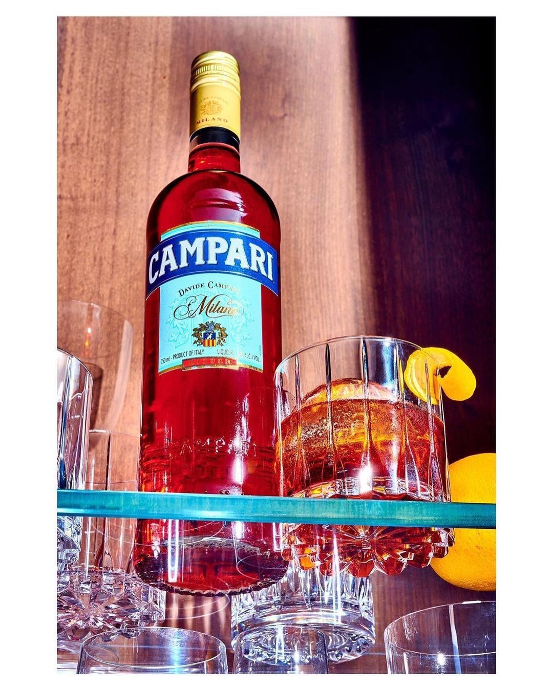 A bottle of Campari and a cocktail sit on a glass shelf, surrounded by empty crystal glasses and orange peel, capturing a bar ambiance.