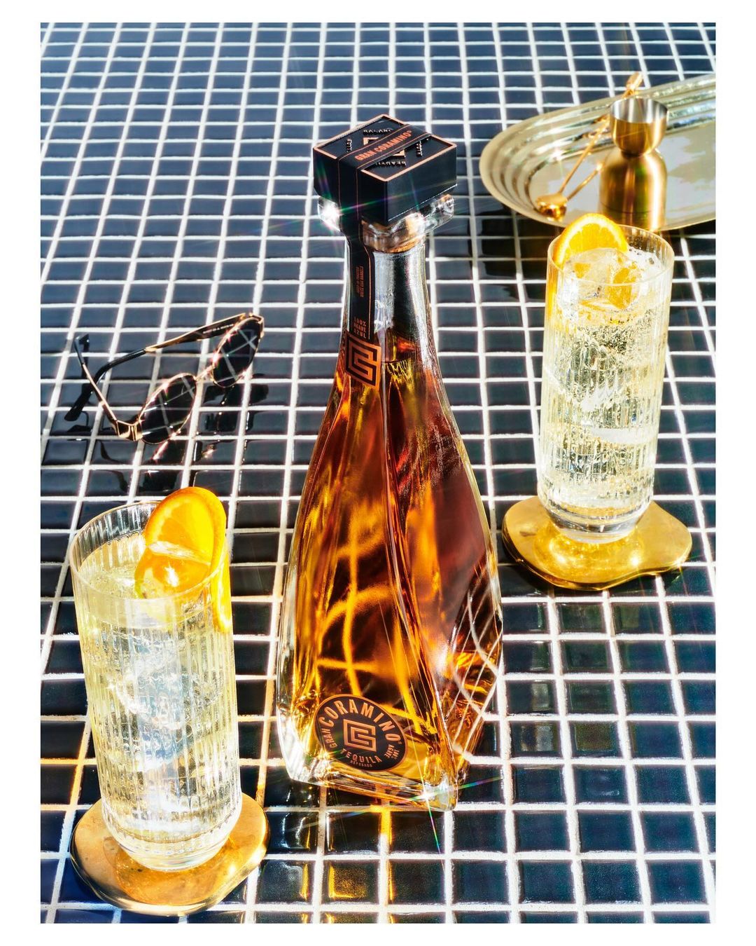 Bottle of Coramino tequila with two glasses of highball cocktails garnished with orange slices, sunglasses and ice on a tile surface.