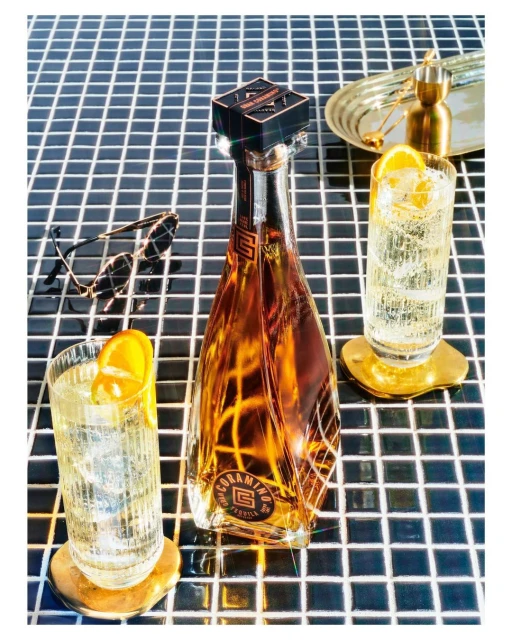 Bottle of Coramino tequila with two glasses of highball cocktails garnished with orange slices, sunglasses and ice on a tile surface.