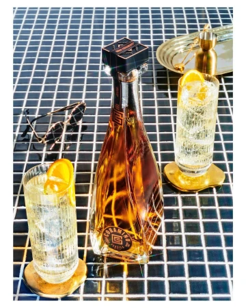 Bottle of Coramino tequila with two glasses of highball cocktails garnished with orange slices, sunglasses and ice on a tile surface.