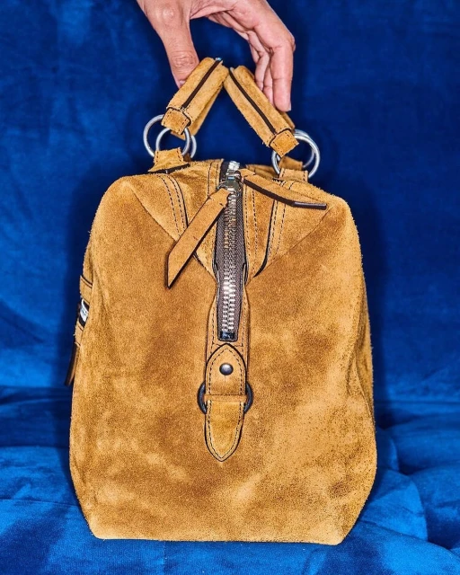 Close-up of a brown suede handbag held up by a fair-skinned hand against a textured blue backdrop, showcasing its details and textures.