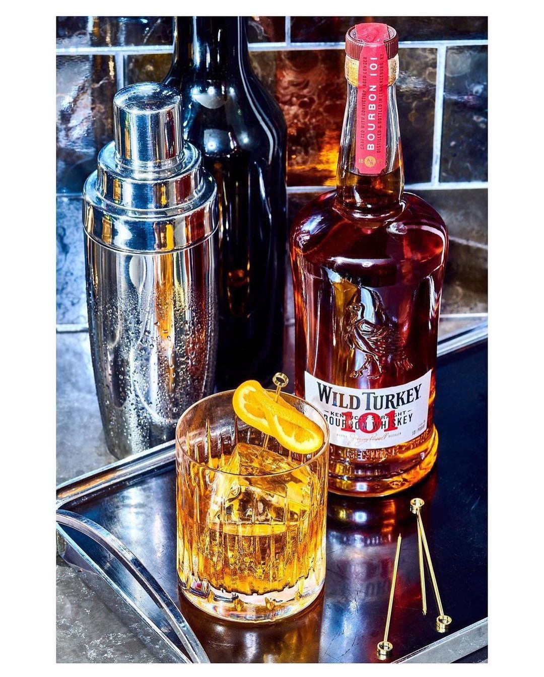 Drink presentation on a silver tray featuring an old-fashioned cocktail with an orange slice, a bottle of Wild Turkey, and a cocktail shaker.