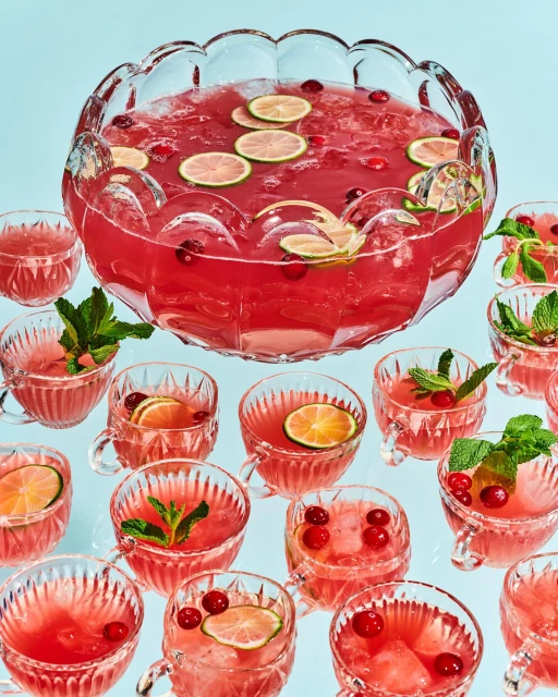 Large clear punch bowl with red punch, garnished with sliced limes, surrounded by smaller punch-filled cups and mint sprigs.