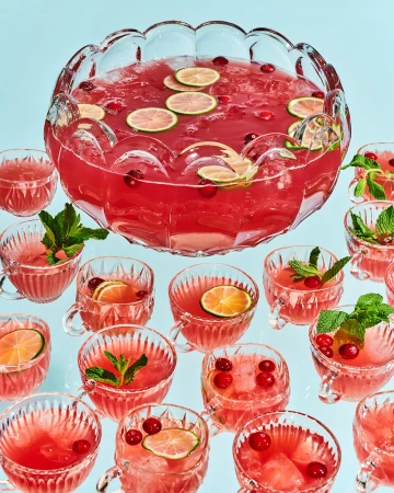 Large clear punch bowl with red punch, garnished with sliced limes, surrounded by smaller punch-filled cups and mint sprigs.