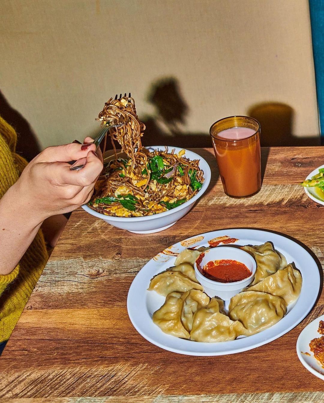 Overhead shot featuring noodle dish, plate of dumplings with dipping sauce, and glass of orange liquid on a wooden table.