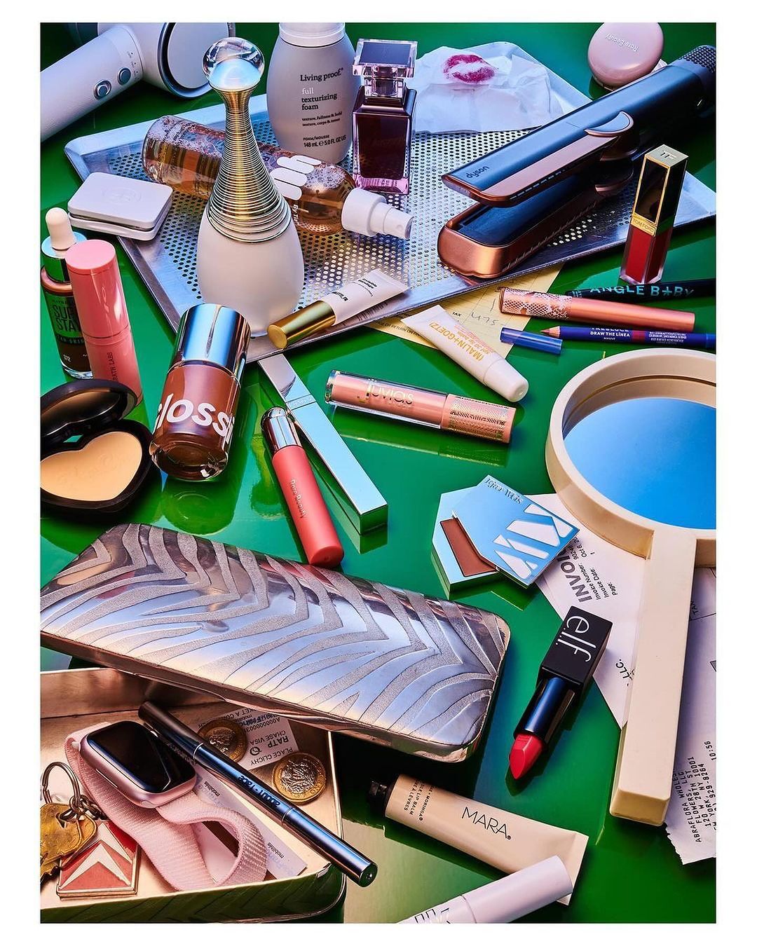 Overhead studio shot of assorted makeup products and beauty accessories, including lipsticks, gloss, foundation, and brushes on green table.