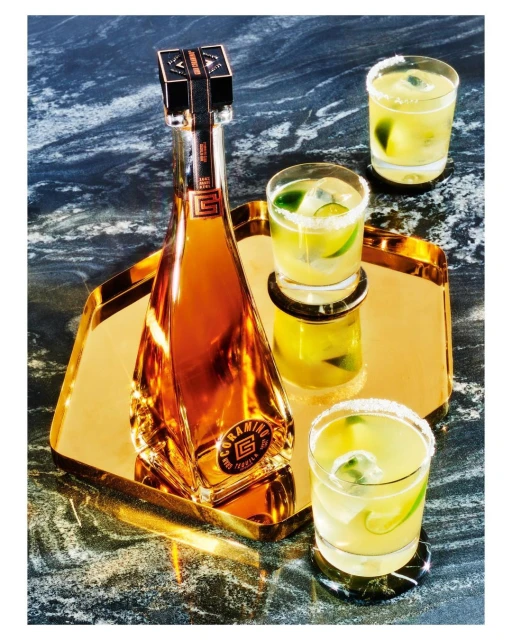 Bottle of Coramino tequila and three margarita cocktails, garnished with lime and salt, on a gold mirrored tray and marble surface.