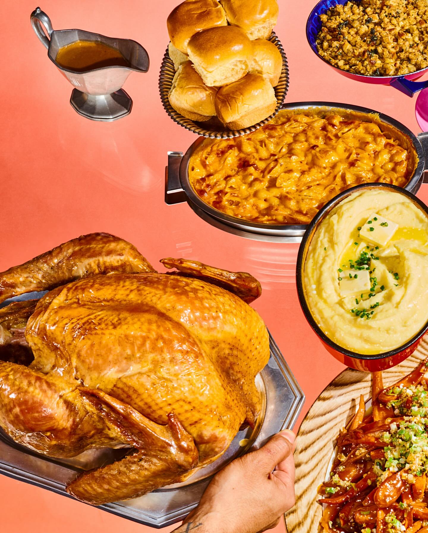Overhead shot of a Thanksgiving feast featuring a turkey, rolls, mac and cheese, sweet potatoes, and mashed potatoes on a pink backdrop.