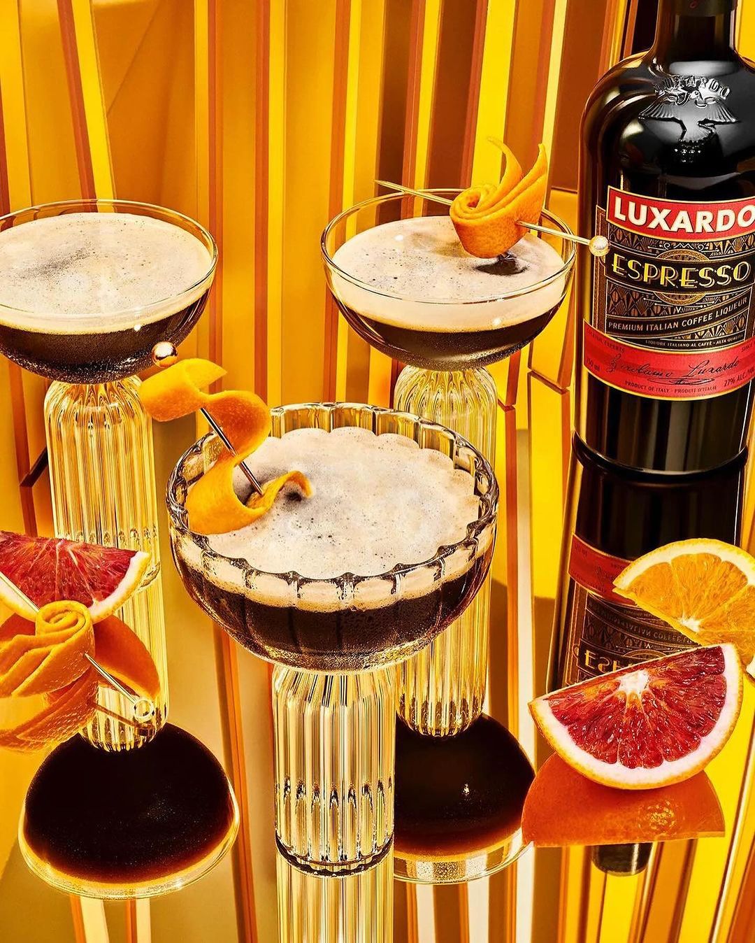 Three espresso martinis topped with orange peel swirls stand on fluted glasses, alongside citrus slices and Luxardo Espresso liqueur.