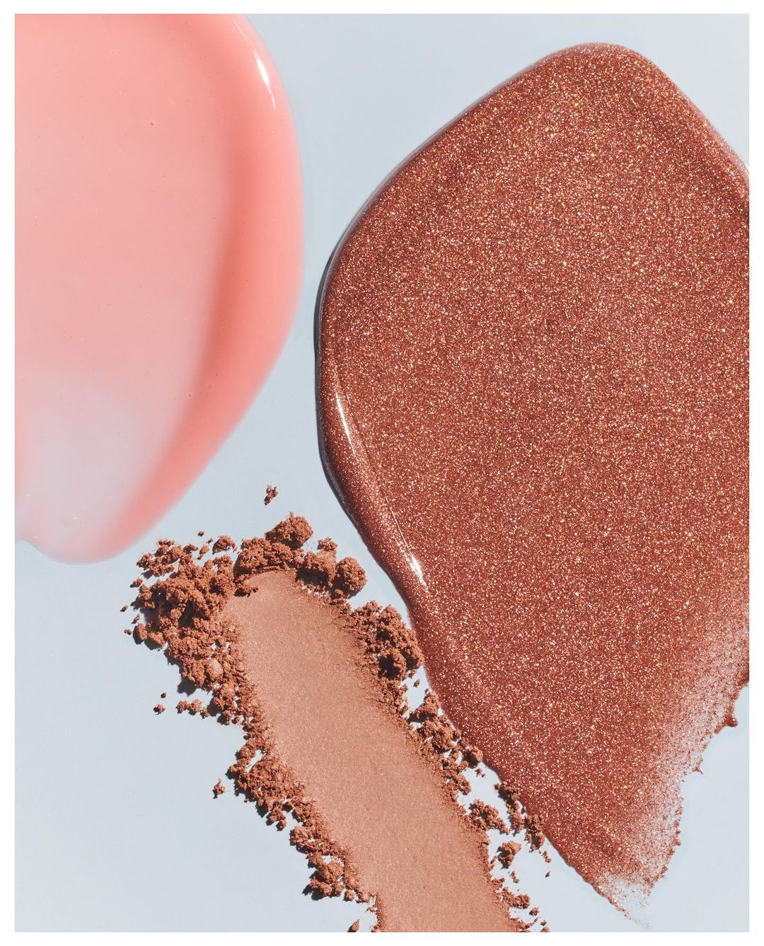 Cosmetic product swatches: glossy pink lip gloss, shimmery bronze highlighter, and neutral blush on a light blue background.