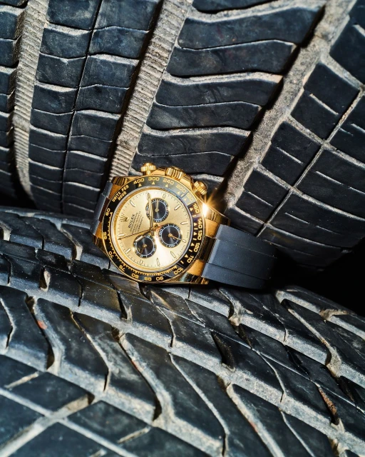 Close-up of a gold chronograph wristwatch with a black band placed on top of a stack of black, ridged automobile tires.