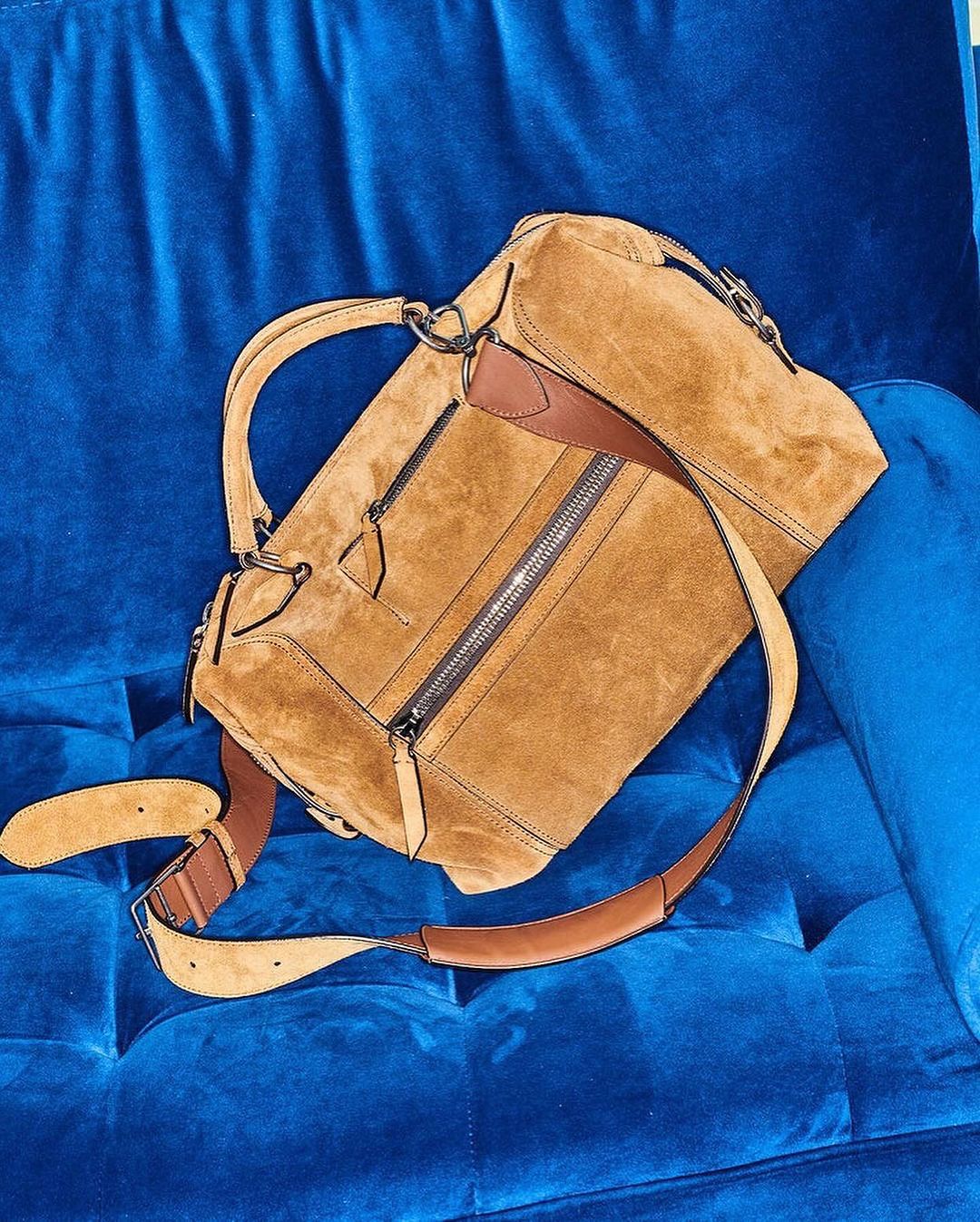 Tan suede satchel handbag with brown leather accents and shoulder strap resting on royal blue velvet tufted furniture.