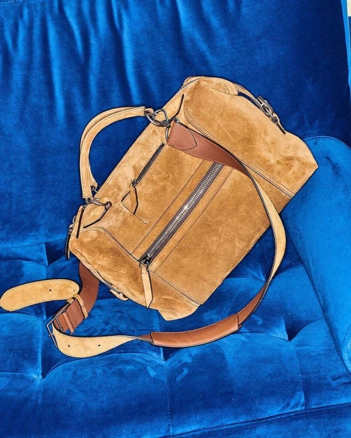 Tan suede satchel handbag with brown leather accents and shoulder strap resting on royal blue velvet tufted furniture.