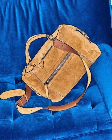Tan suede satchel handbag with brown leather accents and shoulder strap resting on royal blue velvet tufted furniture.