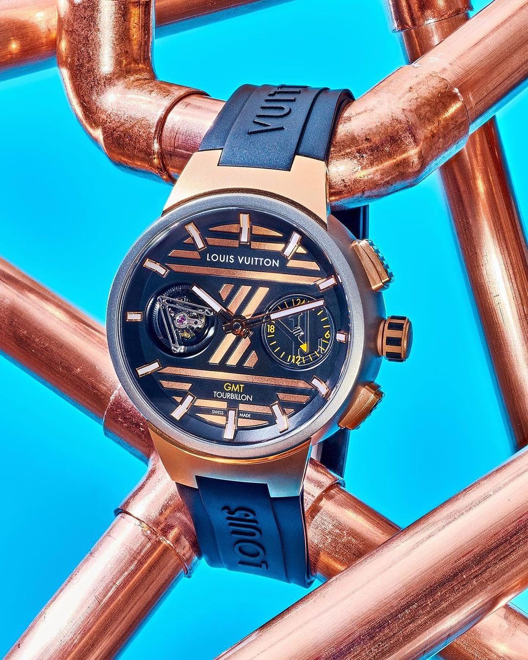 Close-up studio shot of a Louis Vuitton Tambour Moon GMT Tourbillon watch with a black band and rose gold hardware, against a blue background.