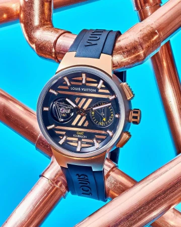 Close-up studio shot of a Louis Vuitton Tambour Moon GMT Tourbillon watch with a black band and rose gold hardware, against a blue background.