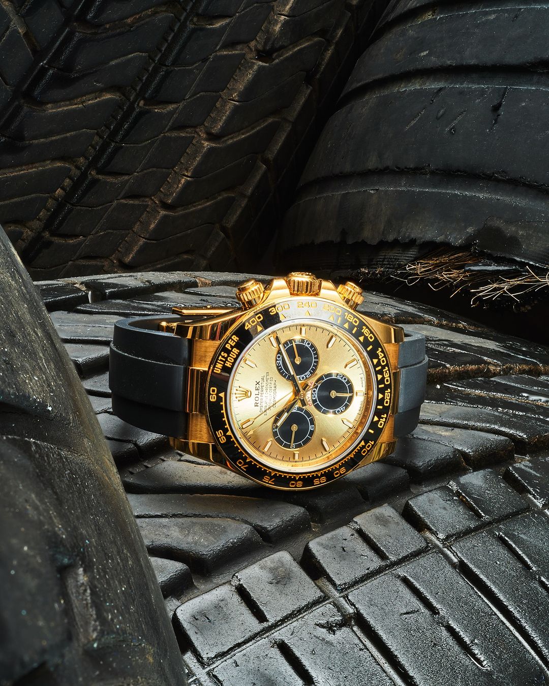 Gold Rolex Daytona wristwatch with a black band sits on a stack of dark, textured tires in a close-up, eye-level product shot.