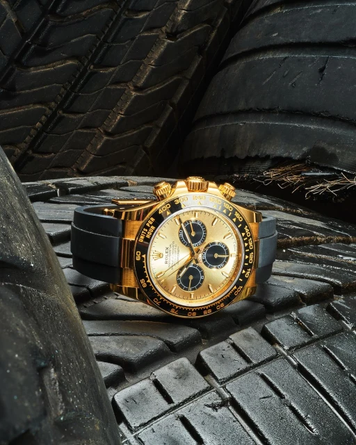 Gold Rolex Daytona wristwatch with a black band sits on a stack of dark, textured tires in a close-up, eye-level product shot.