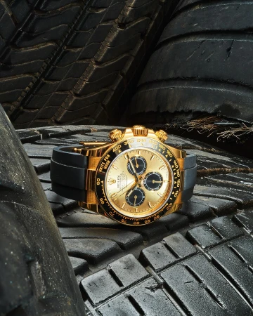 Gold Rolex Daytona wristwatch with a black band sits on a stack of dark, textured tires in a close-up, eye-level product shot.