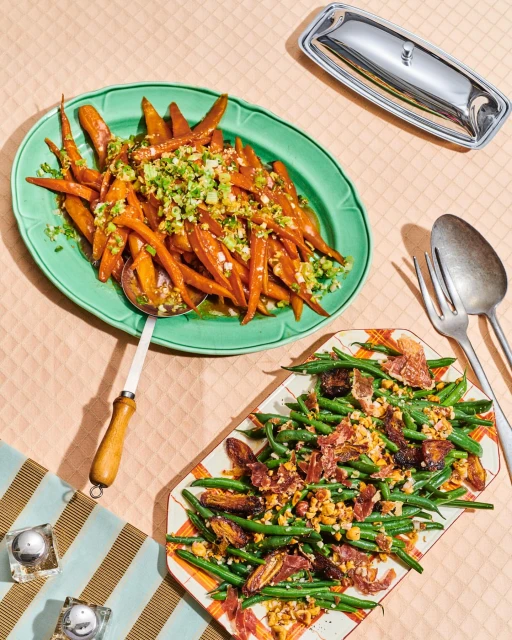 Overhead shot of two platters: one with glazed carrots and the other with green beans, meat, and nuts on a pink tablecloth.
