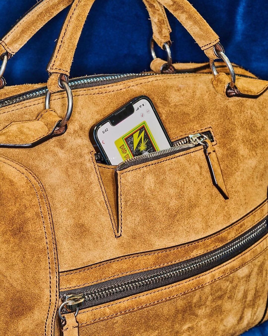 Close-up of a tan suede duffel bag featuring zippers and an iPhone peeking out of its pocket against a dark blue background.