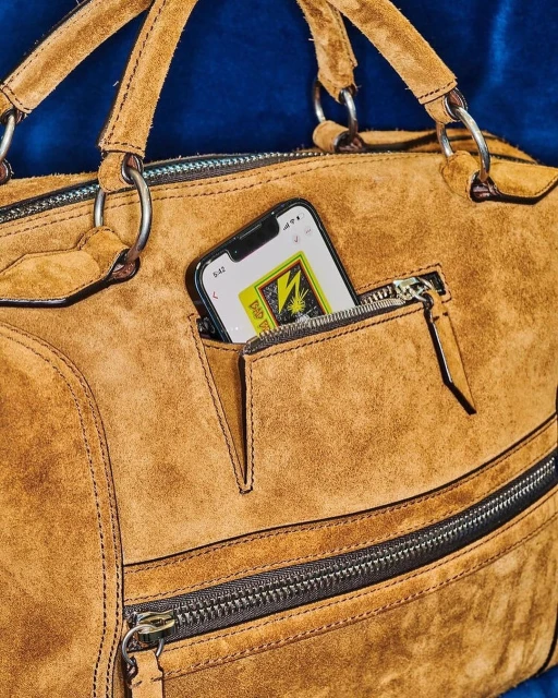 Close-up of a tan suede duffel bag featuring zippers and an iPhone peeking out of its pocket against a dark blue background.