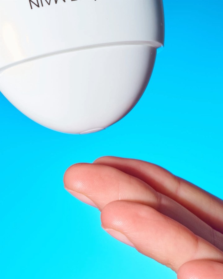 Close-up studio shot of fingers positioned under the nozzle of a white bottle against a vibrant blue background, suggesting skincare or beauty.